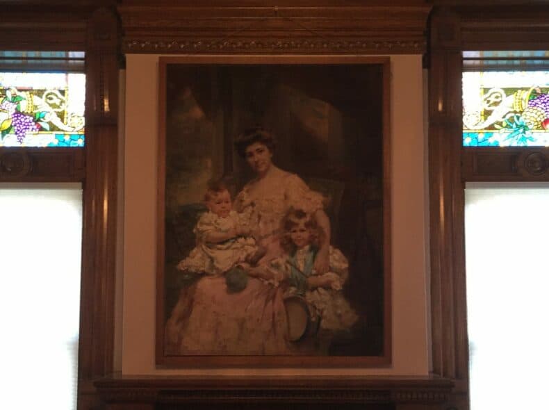 A vintage portrait of a woman seated with two young children, framed in an ornate setting with stained glass windows.
