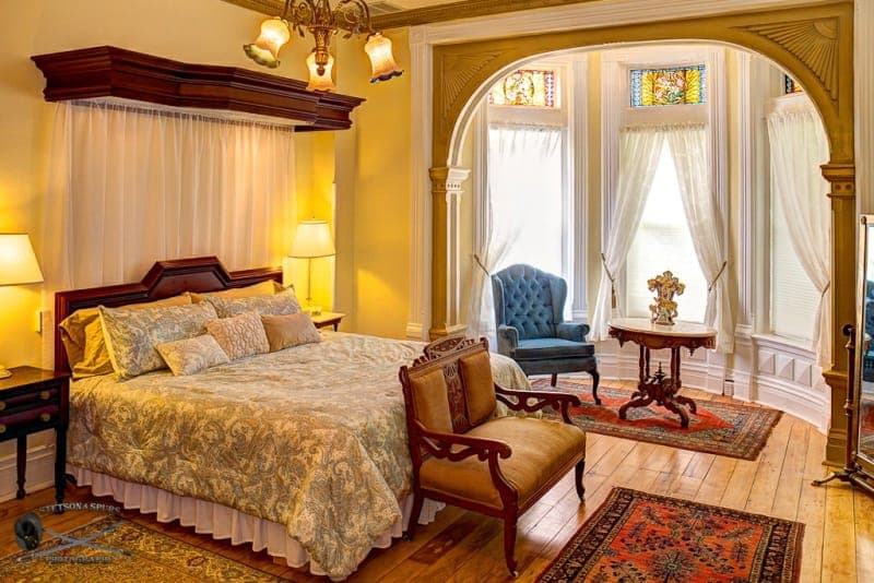 A cozy bedroom featuring a plush bed, antique furniture, and large windows with soft drapes.