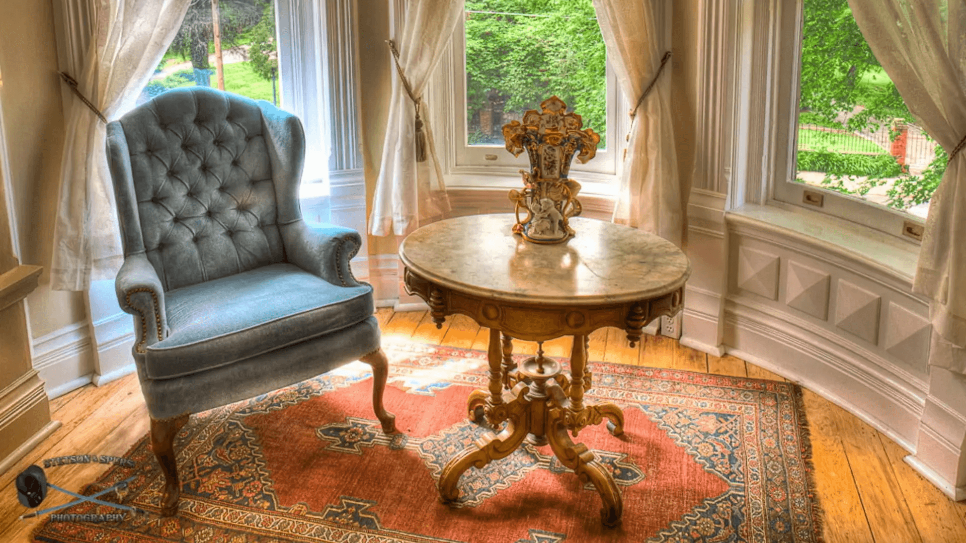 A cozy corner featuring a tufted blue armchair, a round marble-top table with a decorative vase, and sheer curtains.